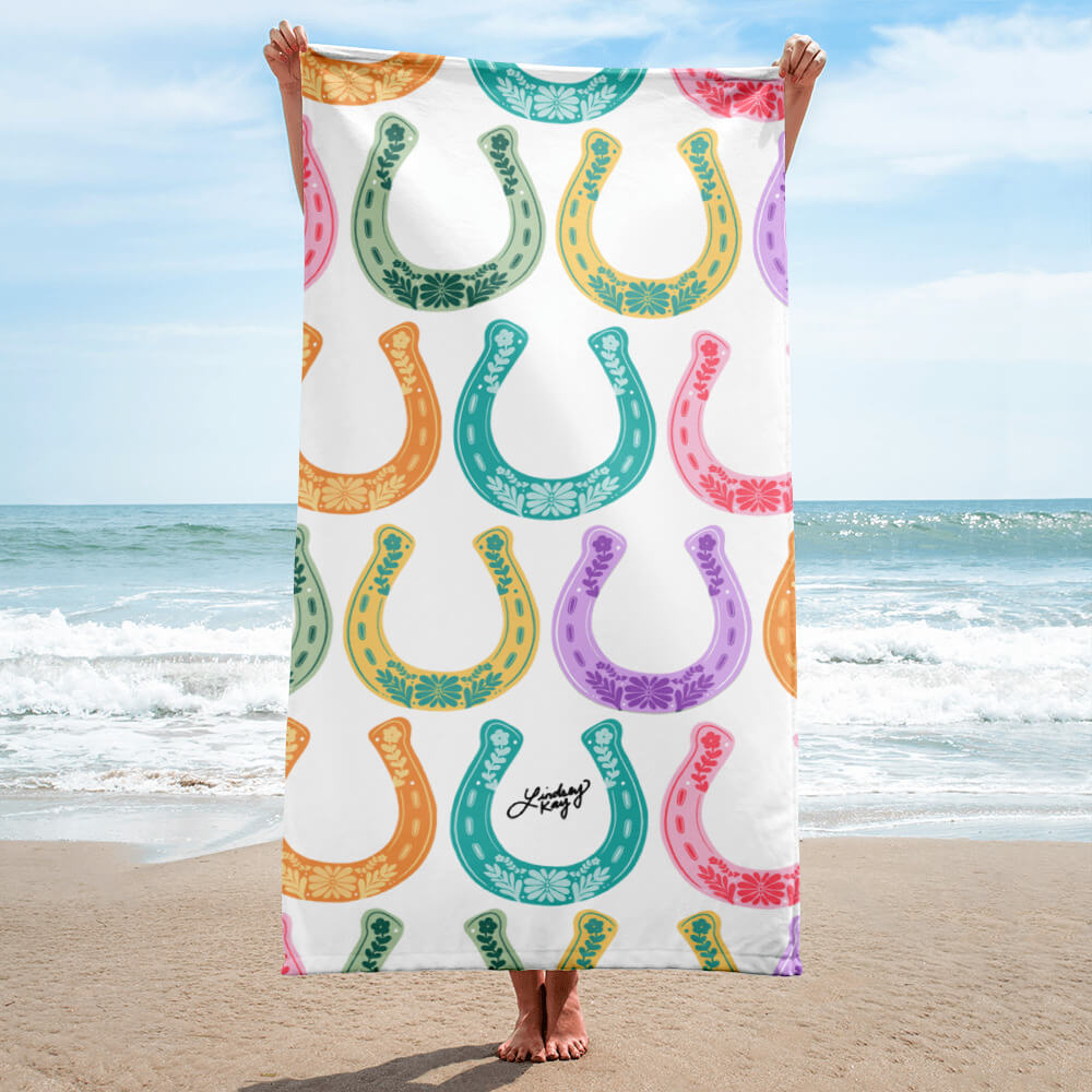Hand illustrated western beach towel by Lindsey Kay Collective. Features bold colorful western-inspired horseshoe design, sized 30×60 inches. Made of soft polyester-cotton blend with absorbent white terry cloth backing. Perfect for beach days, poolside, or summer gifts for cowgirls.