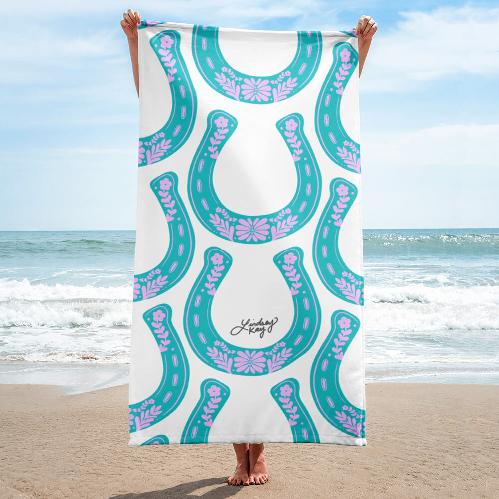 Hand illustrated western beach towel by Lindsey Kay Collective. Features bold turquoise western-inspired horseshoe design, sized 30×60 inches. Made of soft polyester-cotton blend with absorbent white terry cloth backing. Perfect for beach days, poolside, or summer gifts for cowgirls.