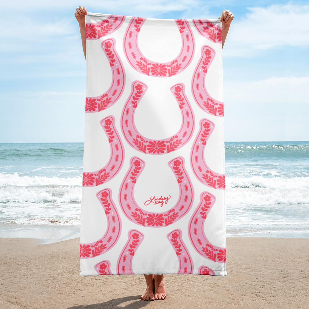 Hand illustrated western beach towel by Lindsey Kay Collective. Features bold pink floral western-inspired horseshoe design, sized 30×60 inches. Made of soft polyester-cotton blend with absorbent white terry cloth backing. Perfect for beach days, poolside, or summer gifts for cowgirls.
